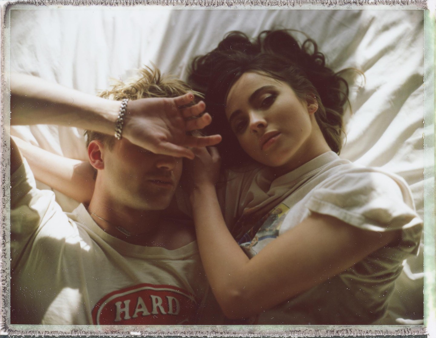 A couple lies on a bed, with the man covering his face with his hand and the woman looking at the viewer.