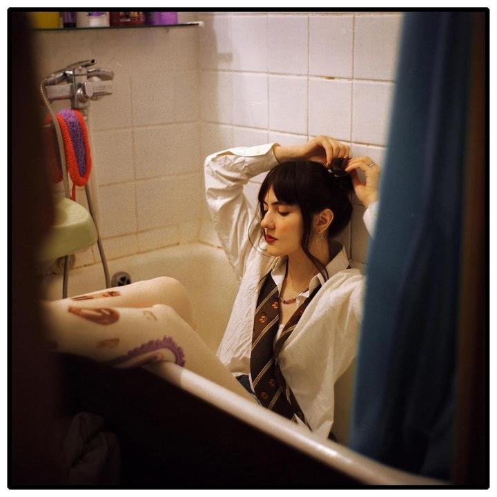 A woman wearing a white shirt and a tie sits in a bathtub and ties her hair.