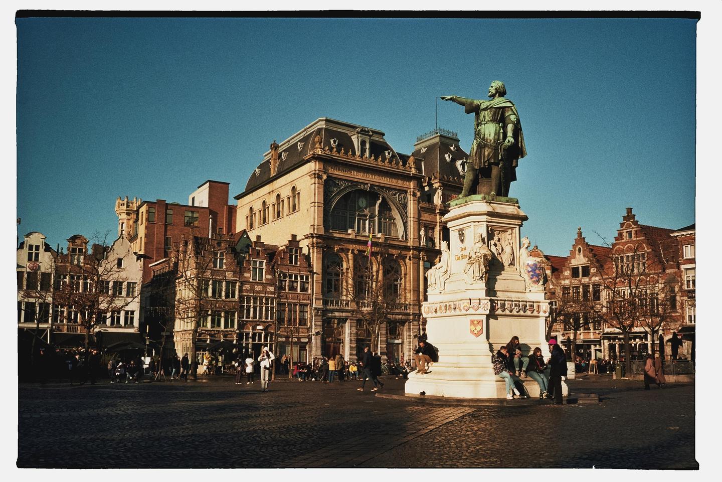 A statue of a man pointing stands in a city square surrounded by historic buildings and people.