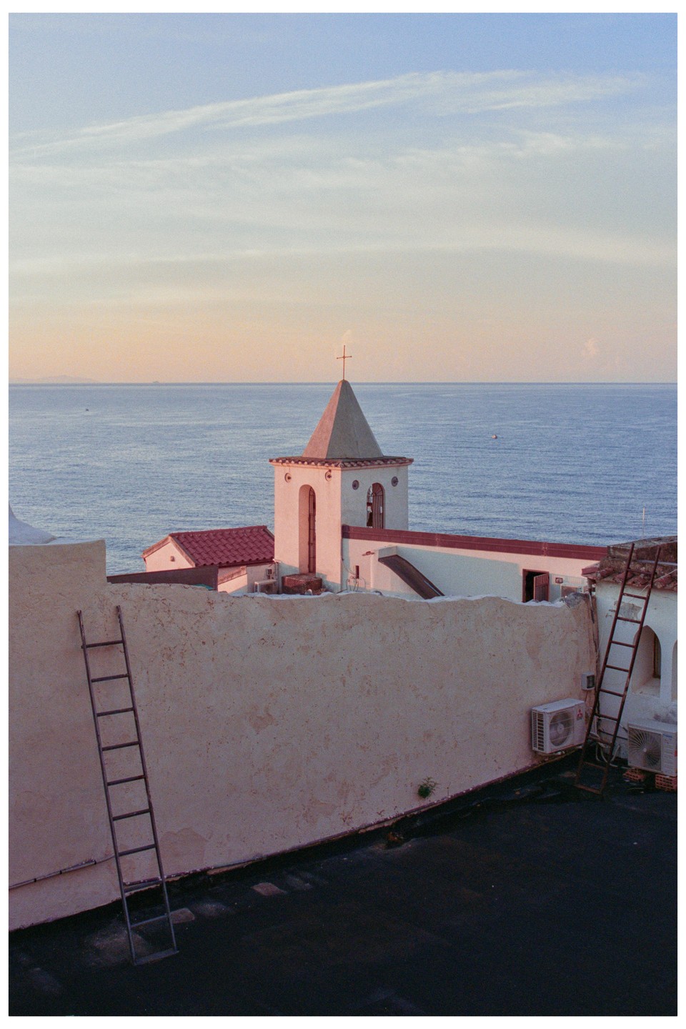 A church tower with a cross on top stands against a vast ocean and sky, with ladders leaning against a wall in the foreground.