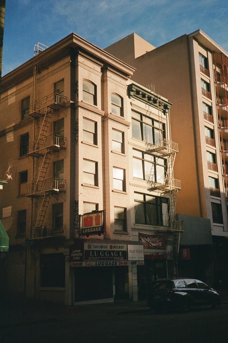 A black car is parked on a street in front of a building with fire escapes and a luggage store.