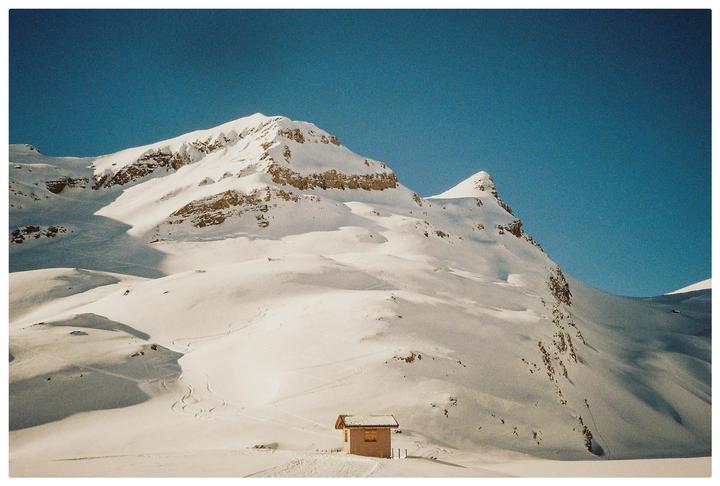 A small cabin sits in a vast, snow-covered mountain landscape under a clear blue sky.
