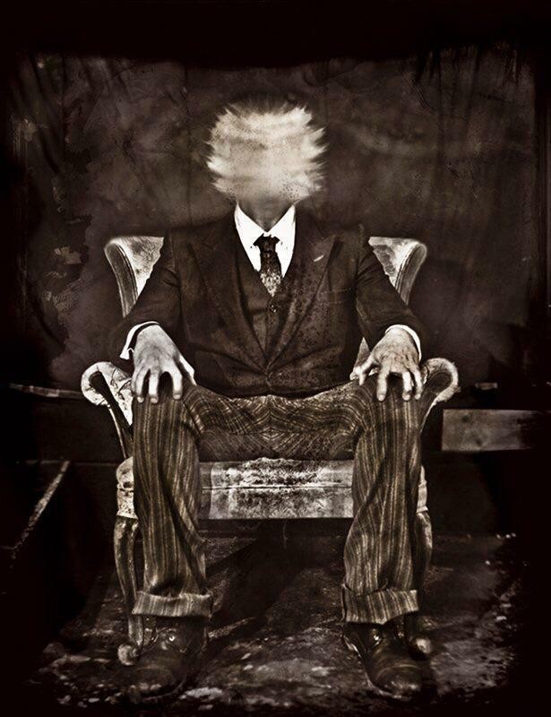 A man in a suit sits in a chair with a blurred, indistinct head.