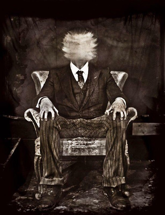 A man in a suit sits in a chair with a blurred, indistinct head.