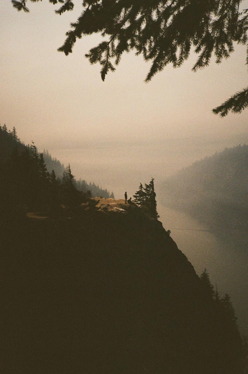 A person stands on a cliff overlooking a hazy valley with a river and forested mountains.
