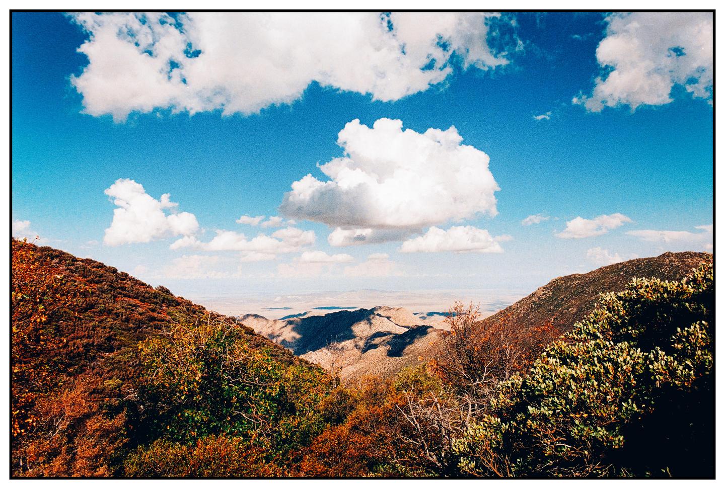 A wide landscape shows a valley with mountains and hills under a blue sky with white clouds.