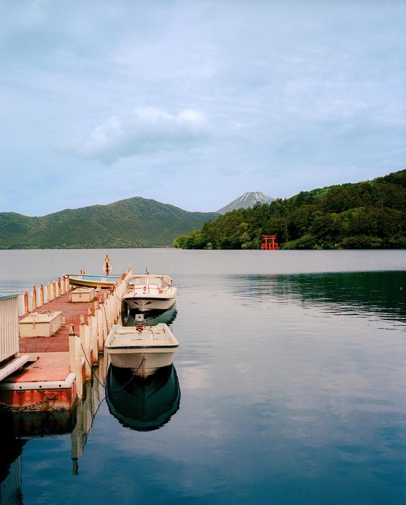Boats are docked at a pier on a lake with a red torii gate and a snow-capped mountain in the distance.