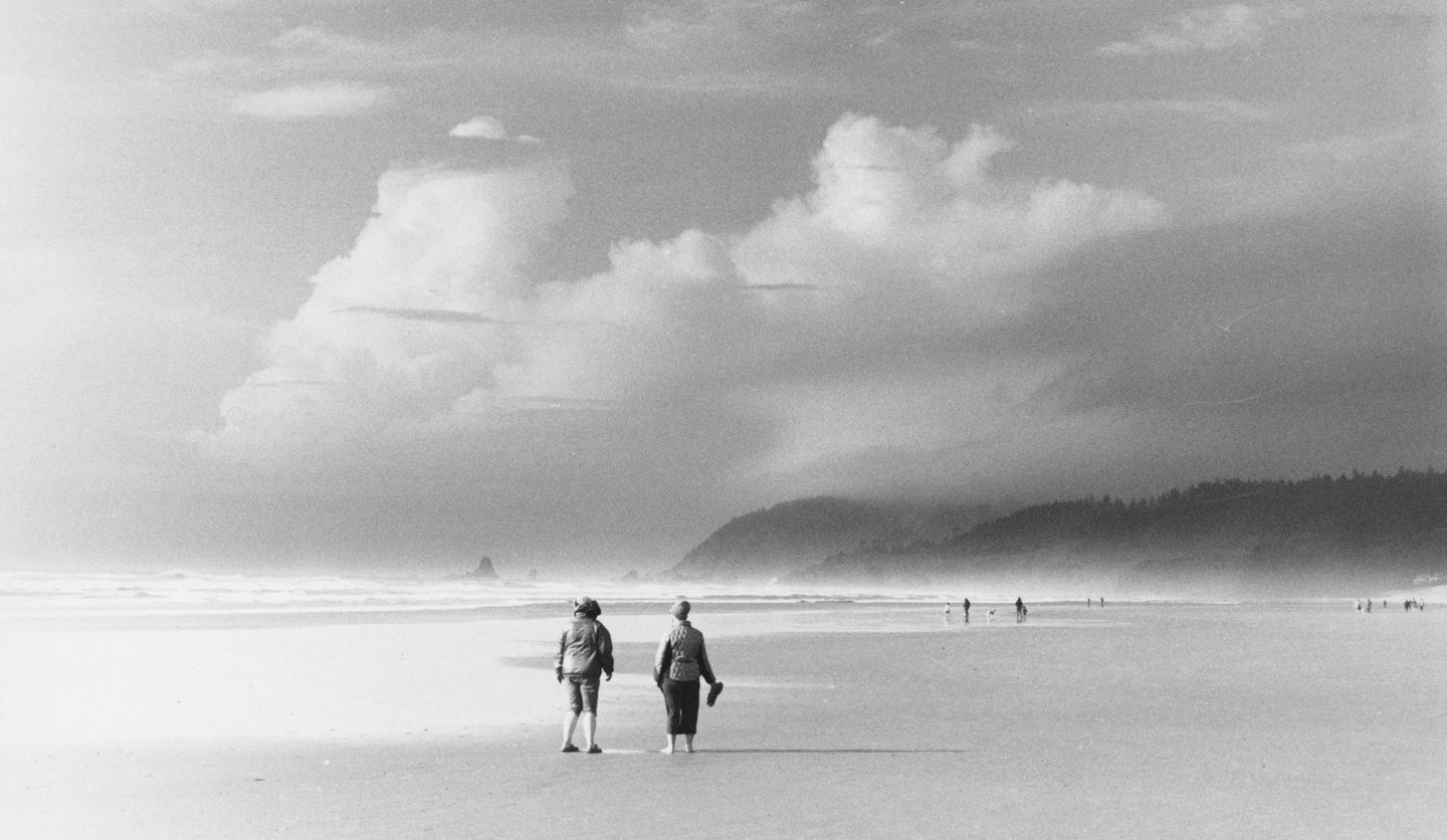 Two women stand on a wide, flat beach looking out at the ocean under a cloudy sky.