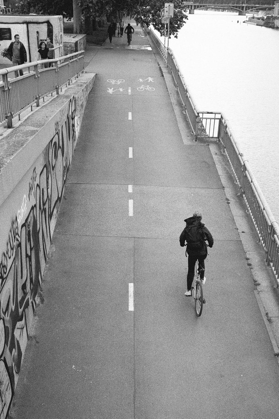 A person rides a bicycle on a path next to a river with a graffiti-covered wall on the left.