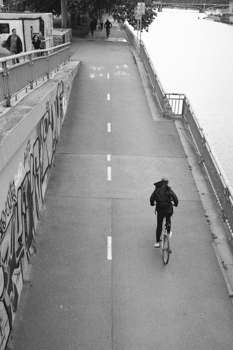 A person rides a bicycle on a path next to a river with a graffiti-covered wall on the left.
