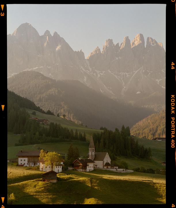 A small village with a church and houses sits in a green valley surrounded by mountains.