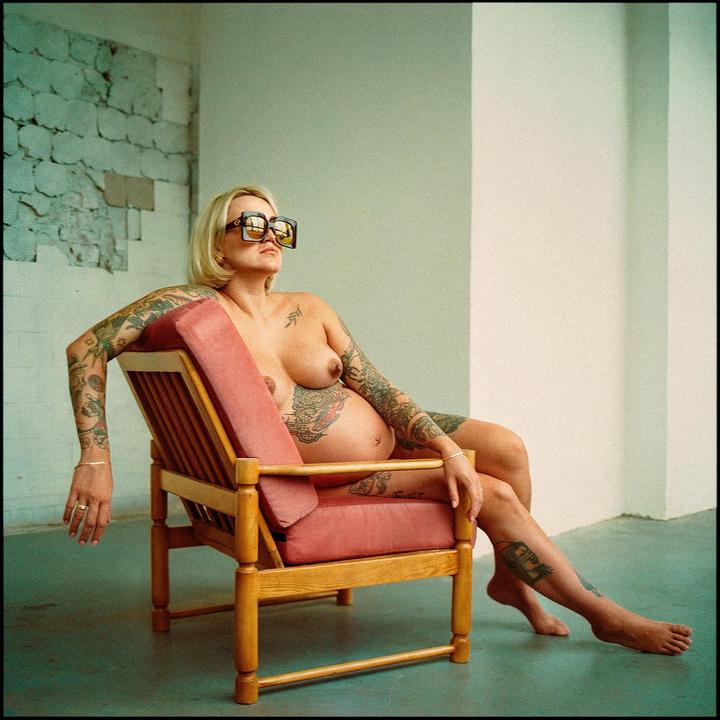 A pregnant woman with tattoos and sunglasses sits in a wooden chair with pink cushions.