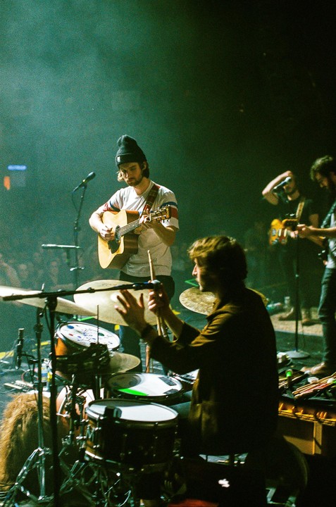A band performs on stage with a guitarist in a beanie singing into a microphone and a drummer in the foreground.