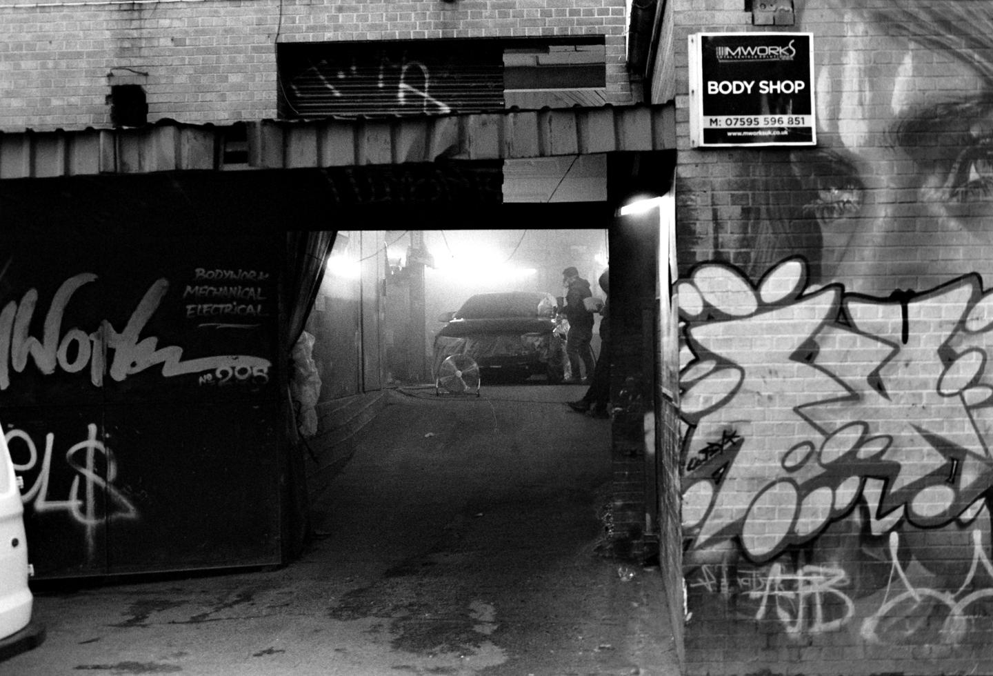 A black and white street photograph shows a car body shop with graffiti on the walls and a car being worked on inside.