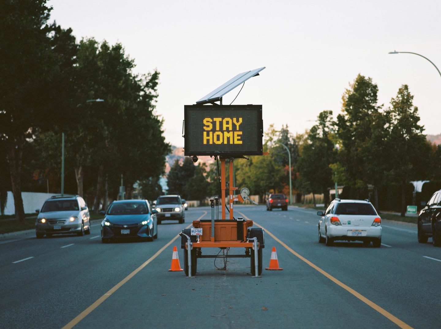 A portable electronic sign displaying "STAY HOME" is set up in the middle of a road with cars driving in both directions.