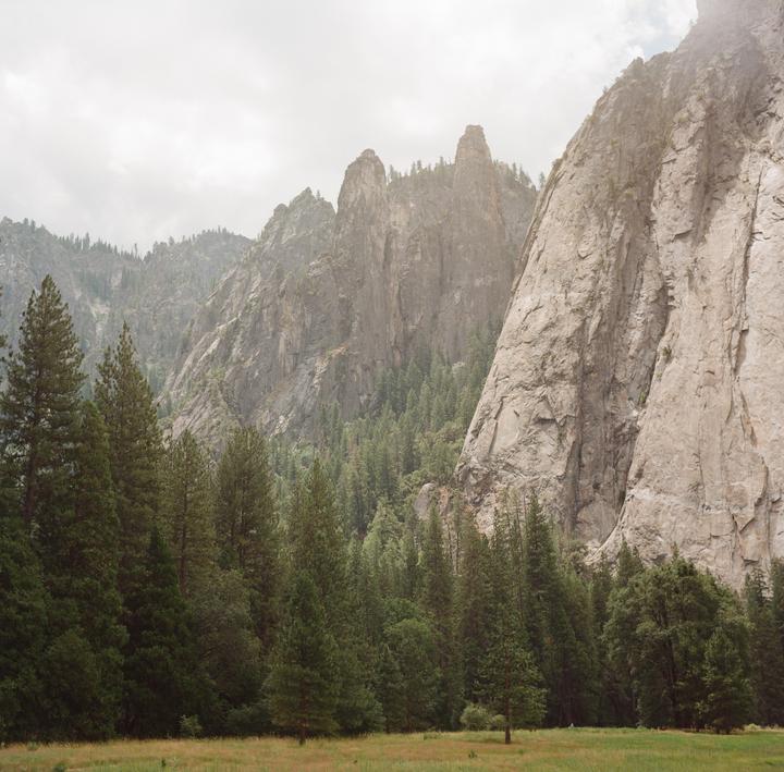 A meadow and forest are at the base of a large mountain range under a hazy sky.