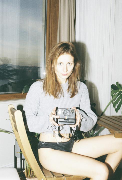 A woman sits on a wooden chair holding an instant camera, with a window and city visible in the background.