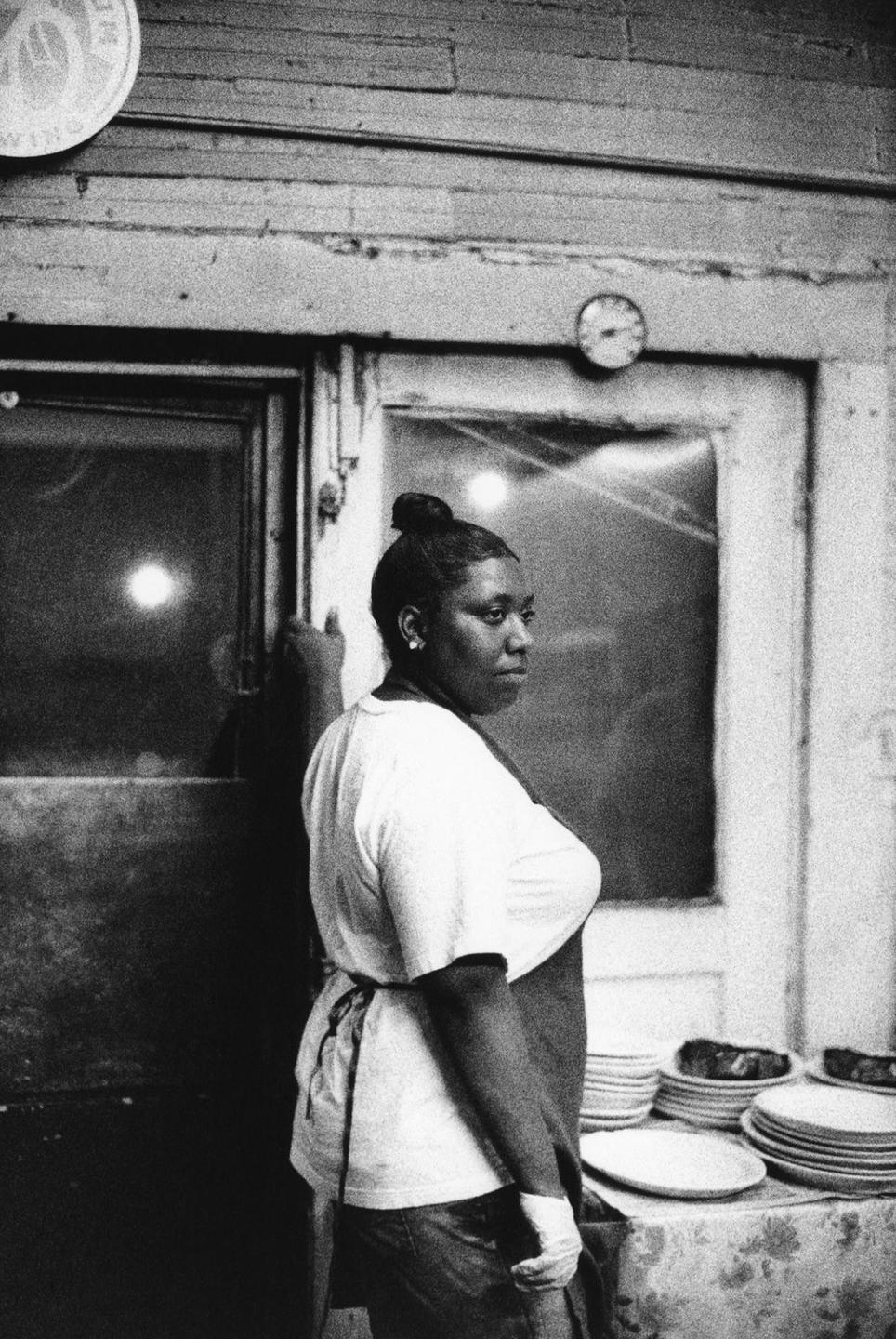 A woman wearing an apron stands next to a table with stacks of plates and food.