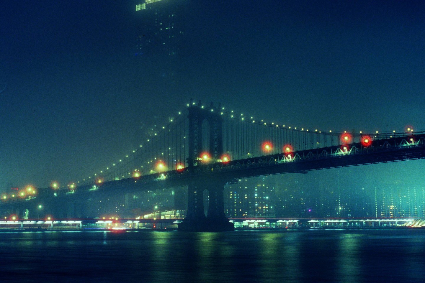 The Manhattan Bridge is illuminated at night, with a hazy sky and city lights in the background.