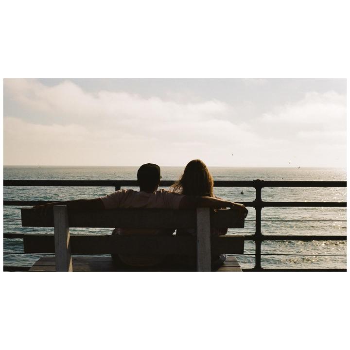A couple sits on a bench overlooking the ocean from a pier.