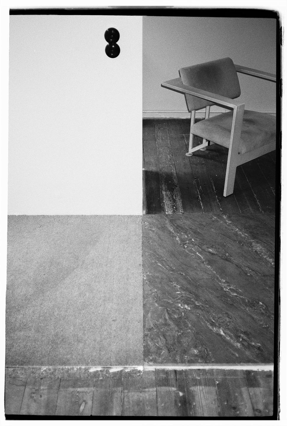 A chair sits on a wooden floor next to a wall with a door handle, with a carpet and a patterned floor in the foreground.