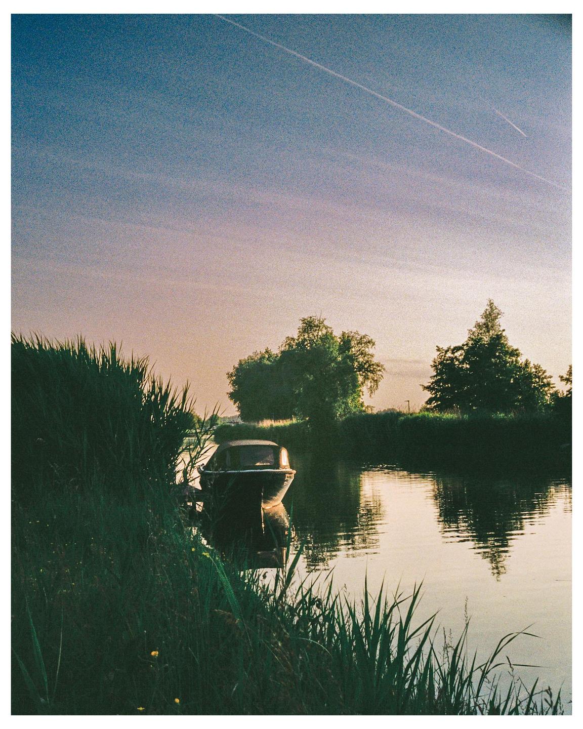A small boat is docked in a river surrounded by tall grass and trees under a blue and orange sky.
