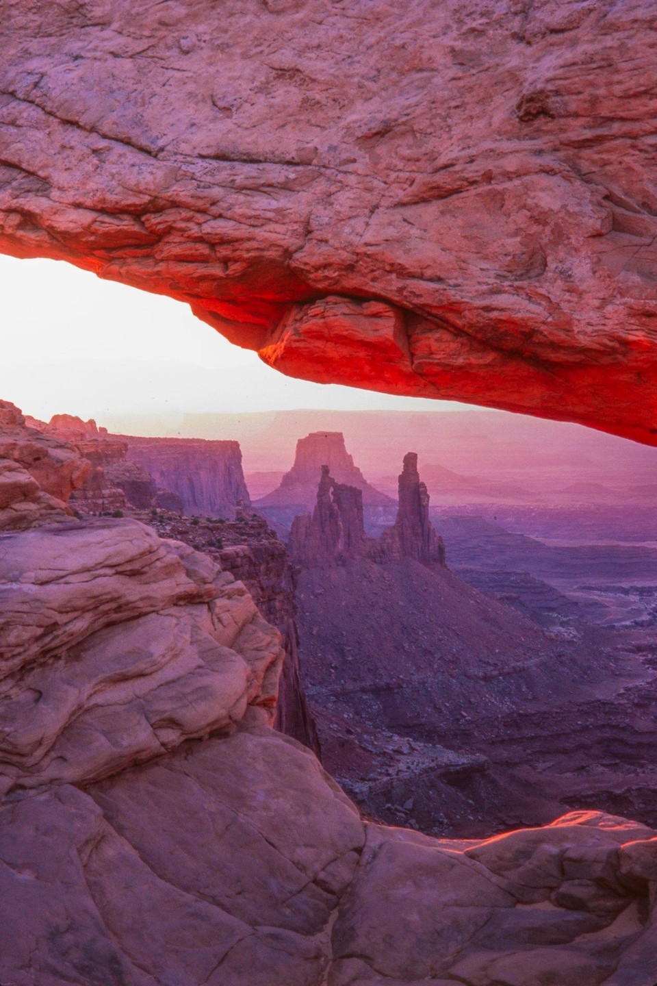 A natural arch frames a desert landscape with rock formations and a vast sky at dawn.