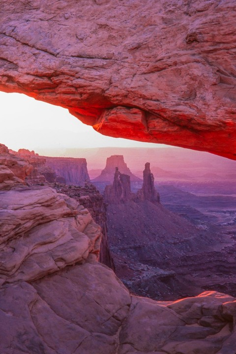 A natural arch frames a desert landscape with rock formations and a vast sky at dawn.