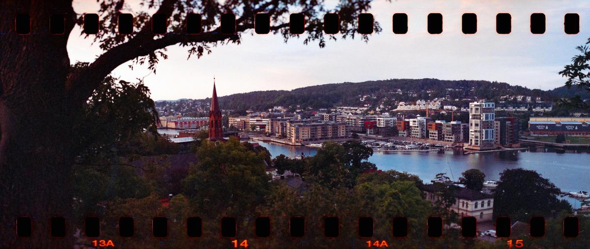 A panoramic view of Tønsberg town shows buildings and a church steeple along a body of water, framed by trees.