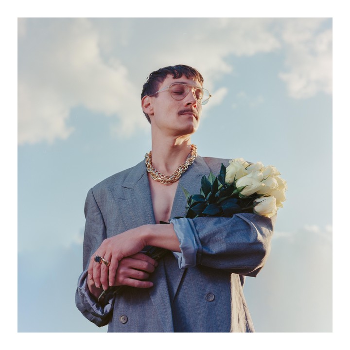 A man wearing a suit jacket and a thick gold chain holds a bouquet of white roses against a cloudy sky.