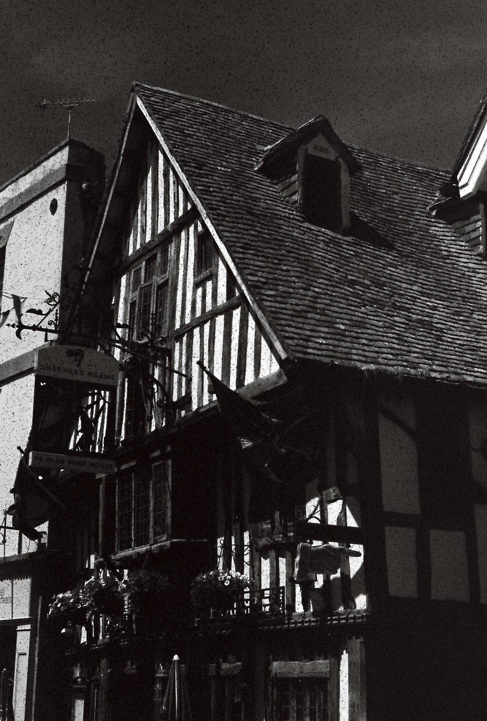 A black and white photo shows a timber-framed building with a steep, tiled roof and a dormer window.