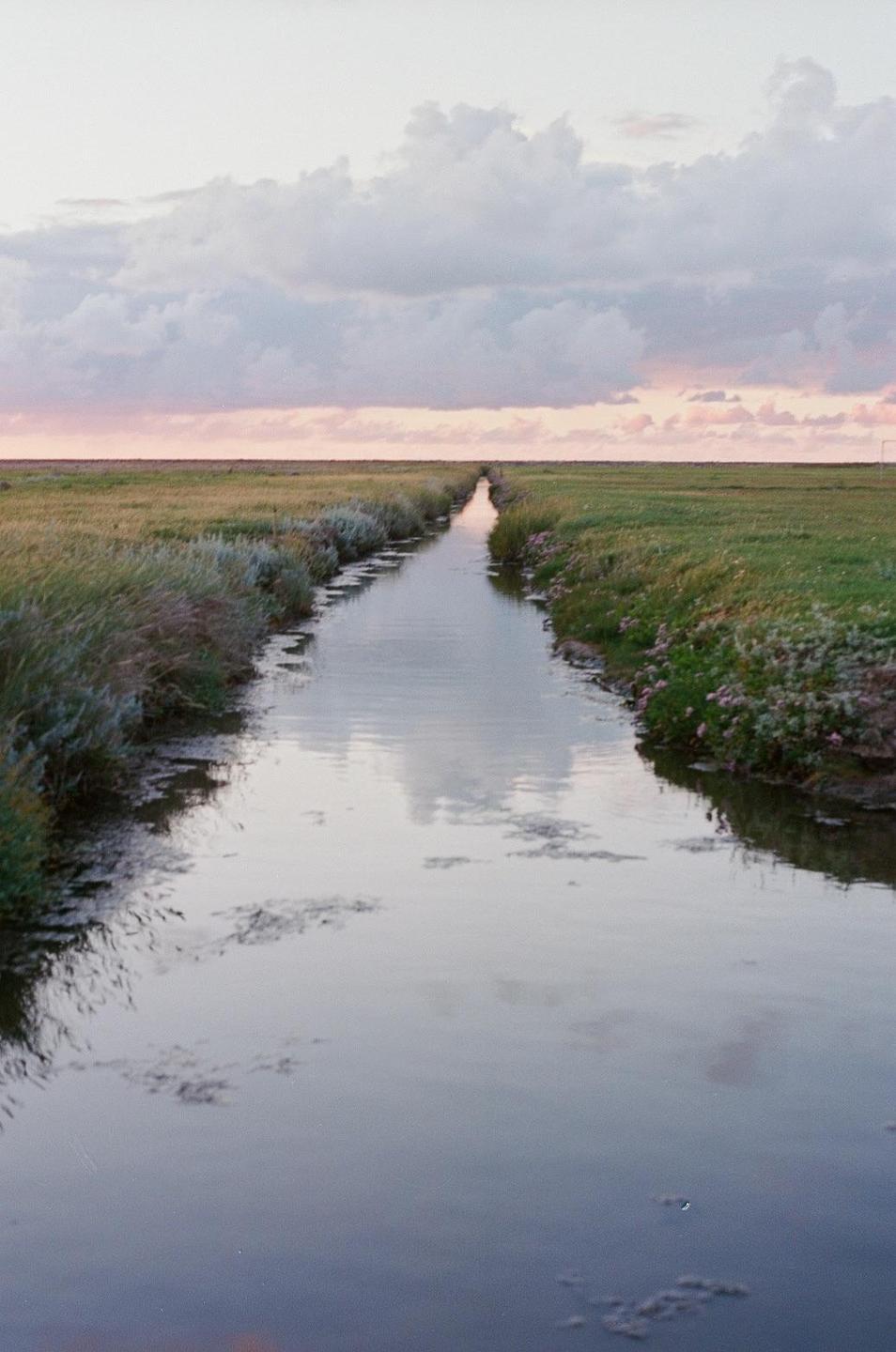 A narrow canal reflects the sky and clouds as it cuts through a grassy field at dusk.