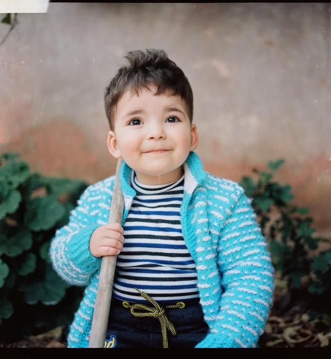 A young boy with dark hair and a blue and white sweater smiles while holding a wooden stick in a garden.