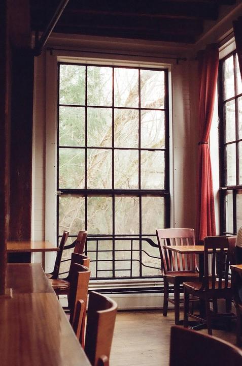 A large window with a grid pattern looks out onto trees, with wooden tables and chairs in the foreground.