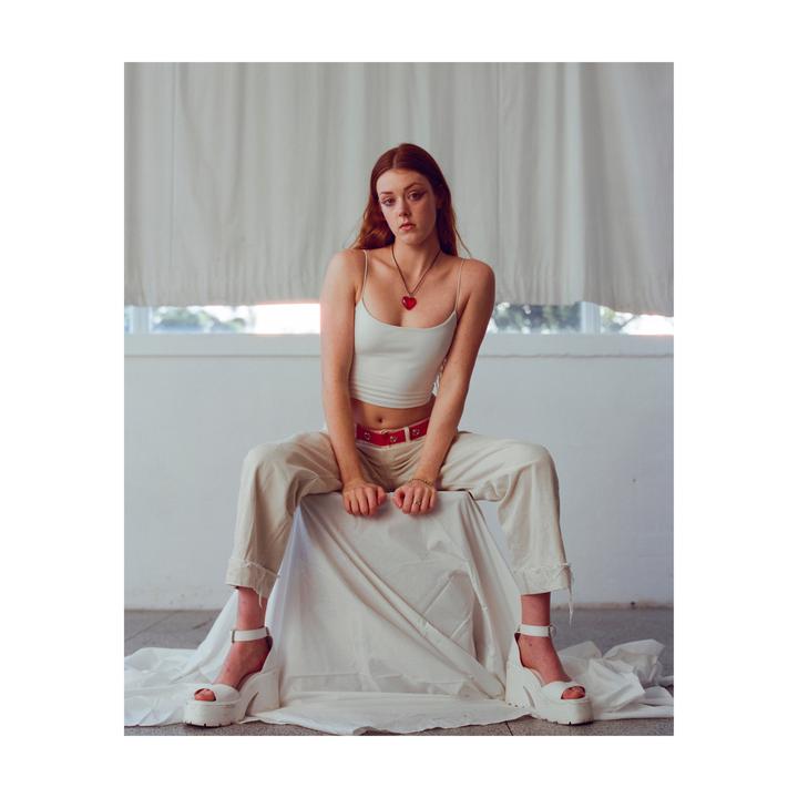 A woman with red hair sits on a draped surface, wearing a white top, light pants, and platform shoes.
