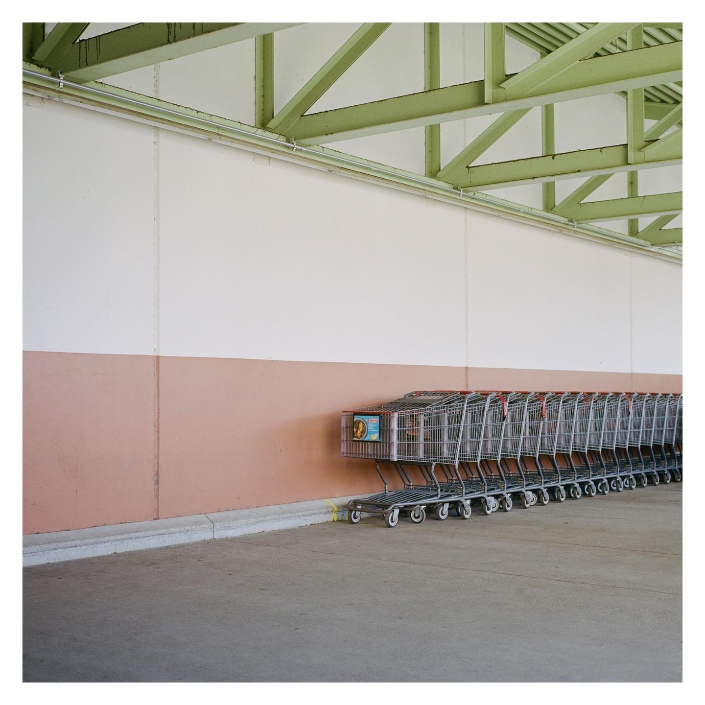A long line of shopping carts is parked against a two-toned wall under a green metal ceiling structure.