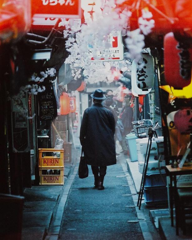 A man in a hat and coat walks down a narrow alley lined with shops and lanterns, with cherry blossoms overhead.