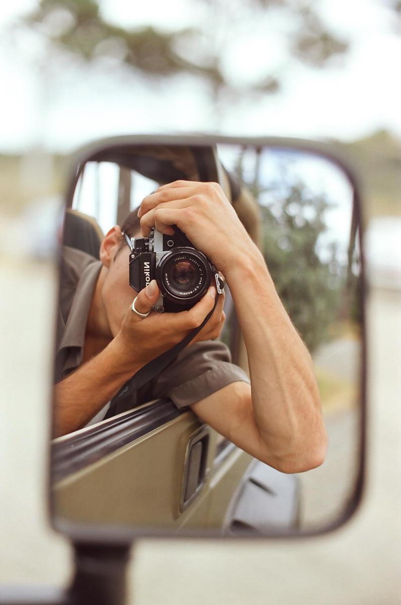 A man is reflected in a side mirror, holding a camera to his eye.