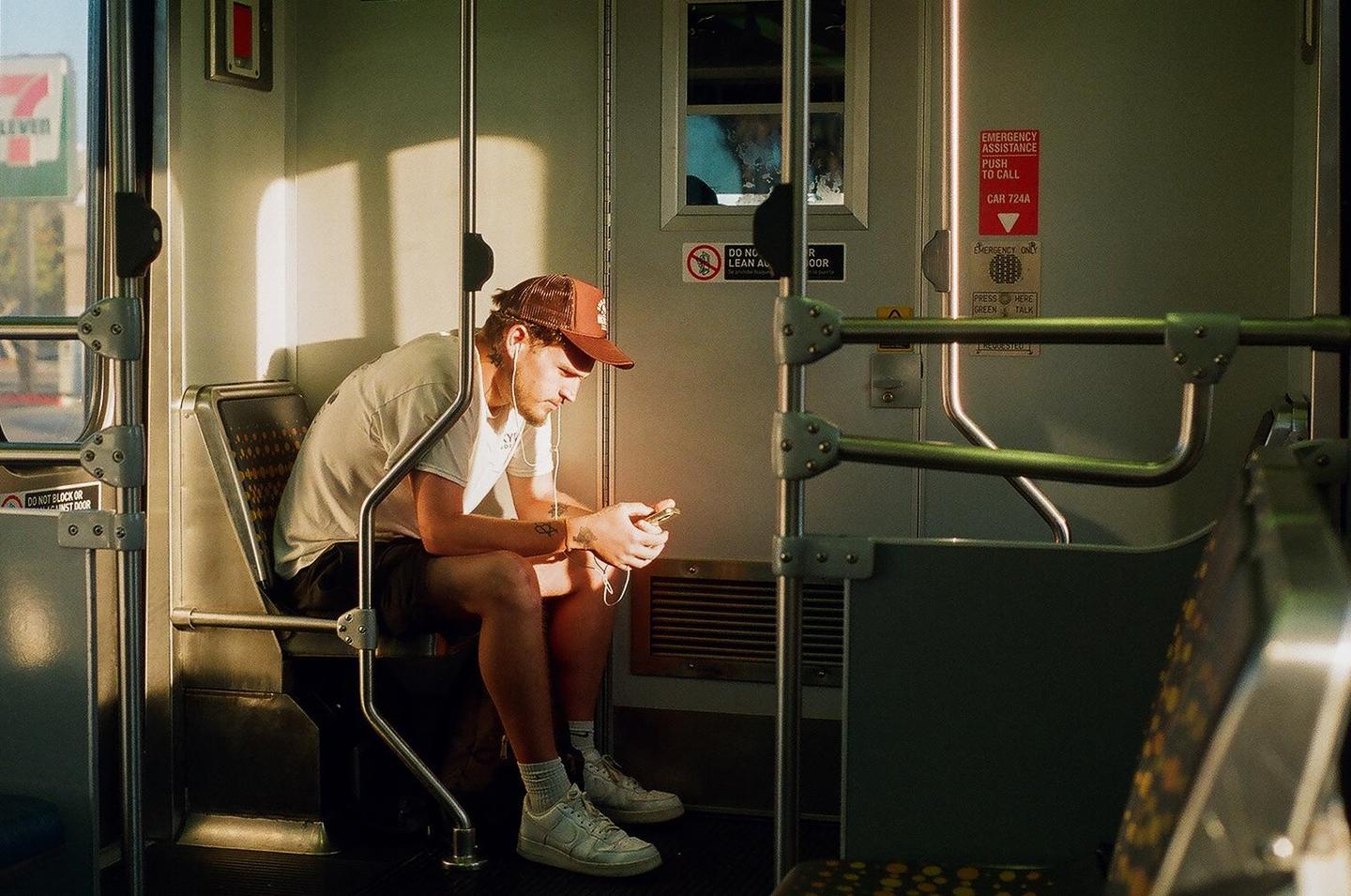 A man wearing a baseball cap and earphones sits on a train, looking at his phone.