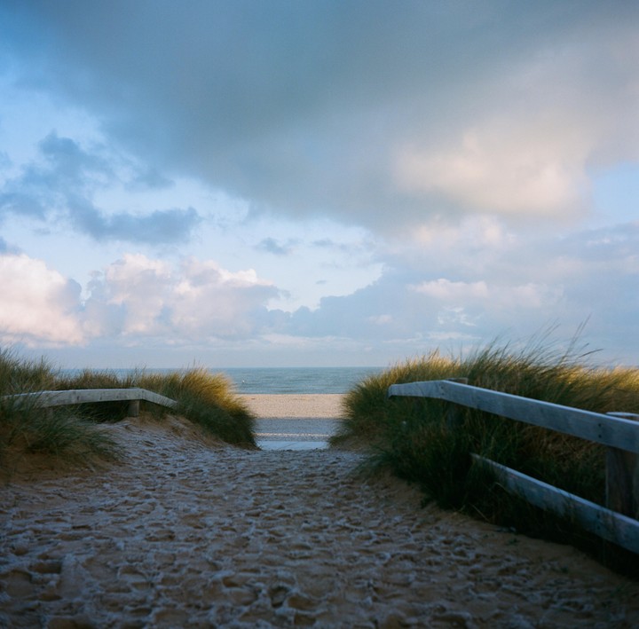 A sandy path leads through grassy dunes to a beach and the ocean under a cloudy sky.