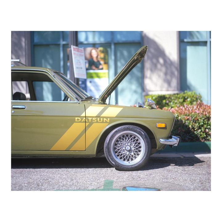 A green Datsun car with its hood open is parked on a street.