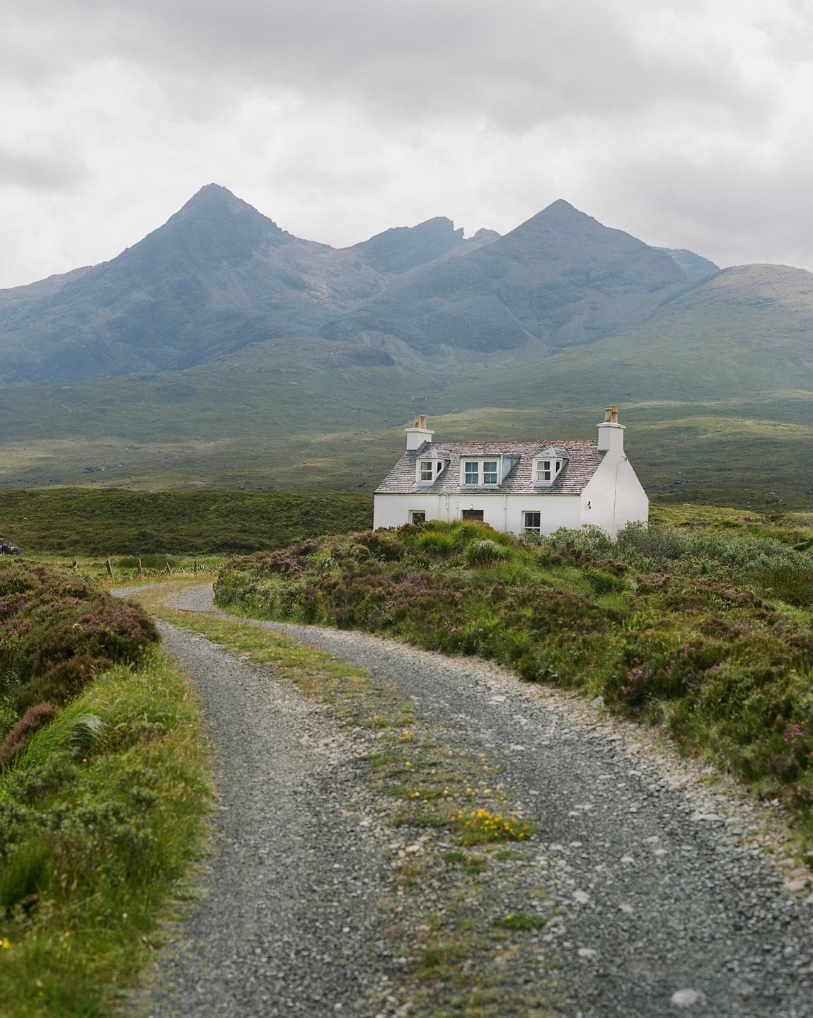 A white house with dormer windows sits in a green and purple landscape with a gravel road leading to it and mountains in the background.