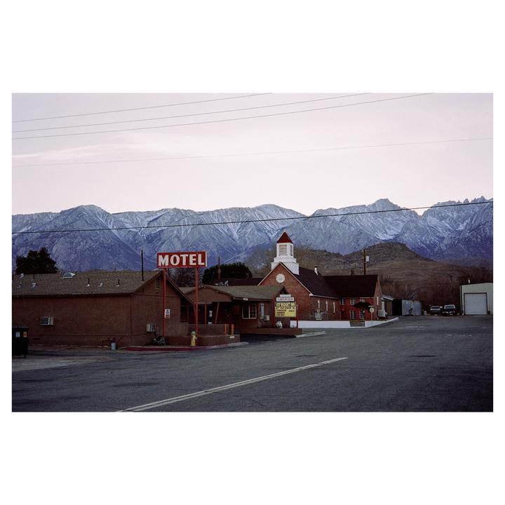 A motel sign stands in front of a brick building with a church-like steeple, with snow-capped mountains in the background.
