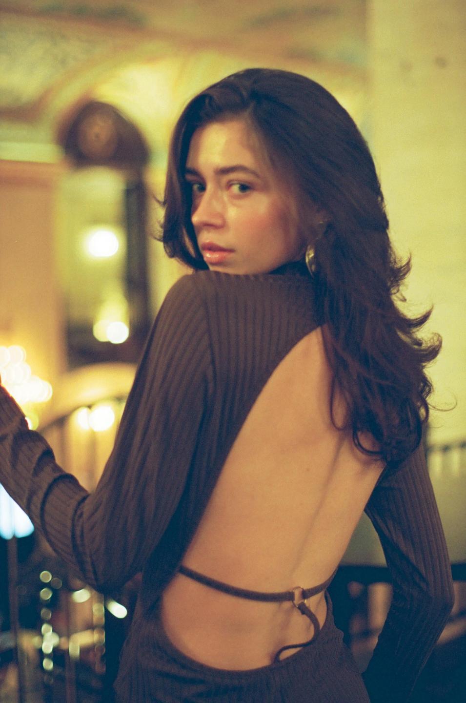 A woman with long hair and an open-back dress looks over her shoulder.