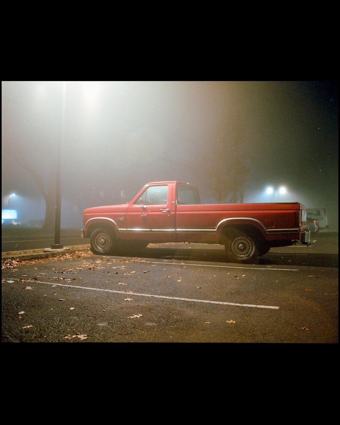 A red pickup truck is parked on an asphalt lot covered in autumn leaves, shrouded in heavy fog at night.