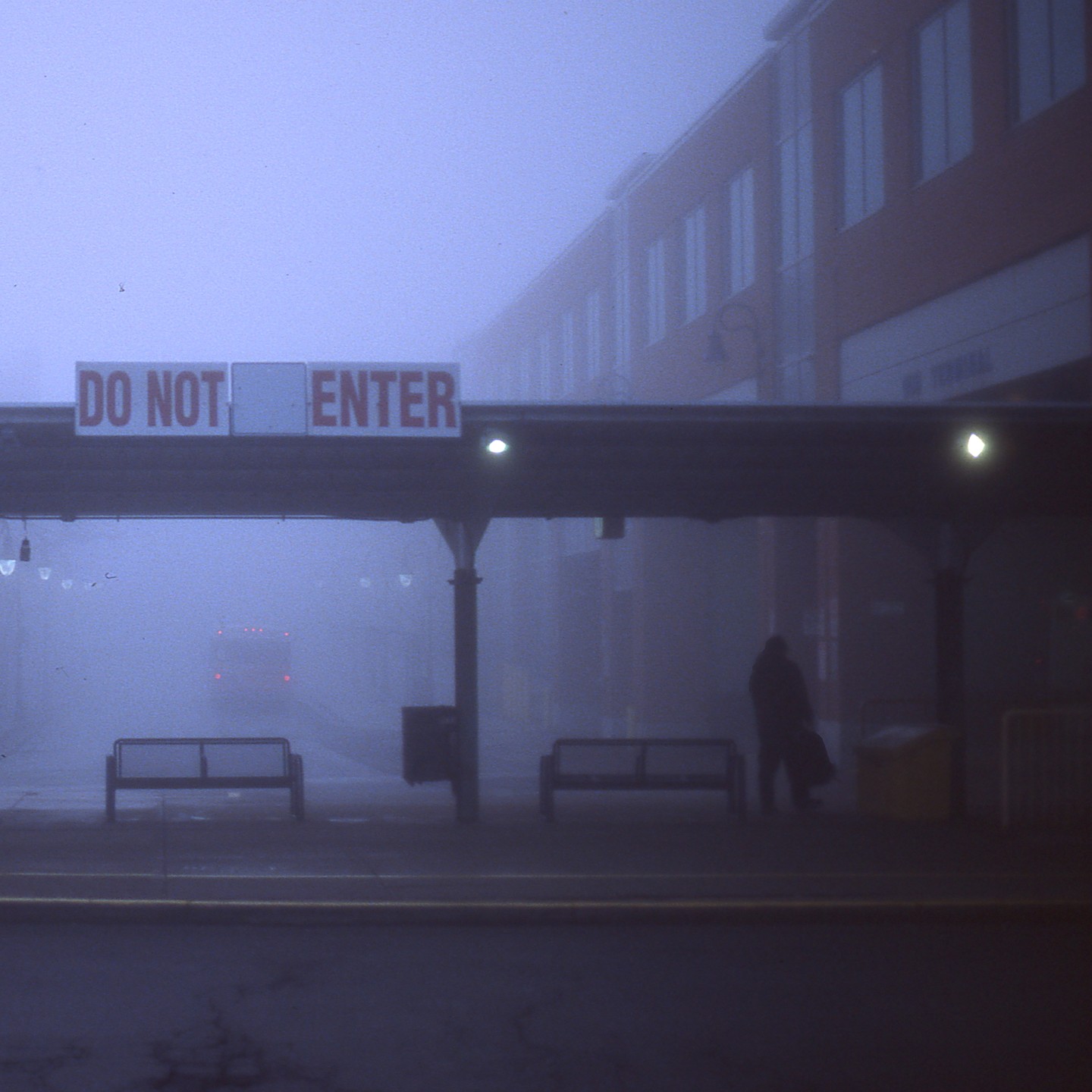 A person stands under a "DO NOT ENTER" sign at a bus terminal in the fog.