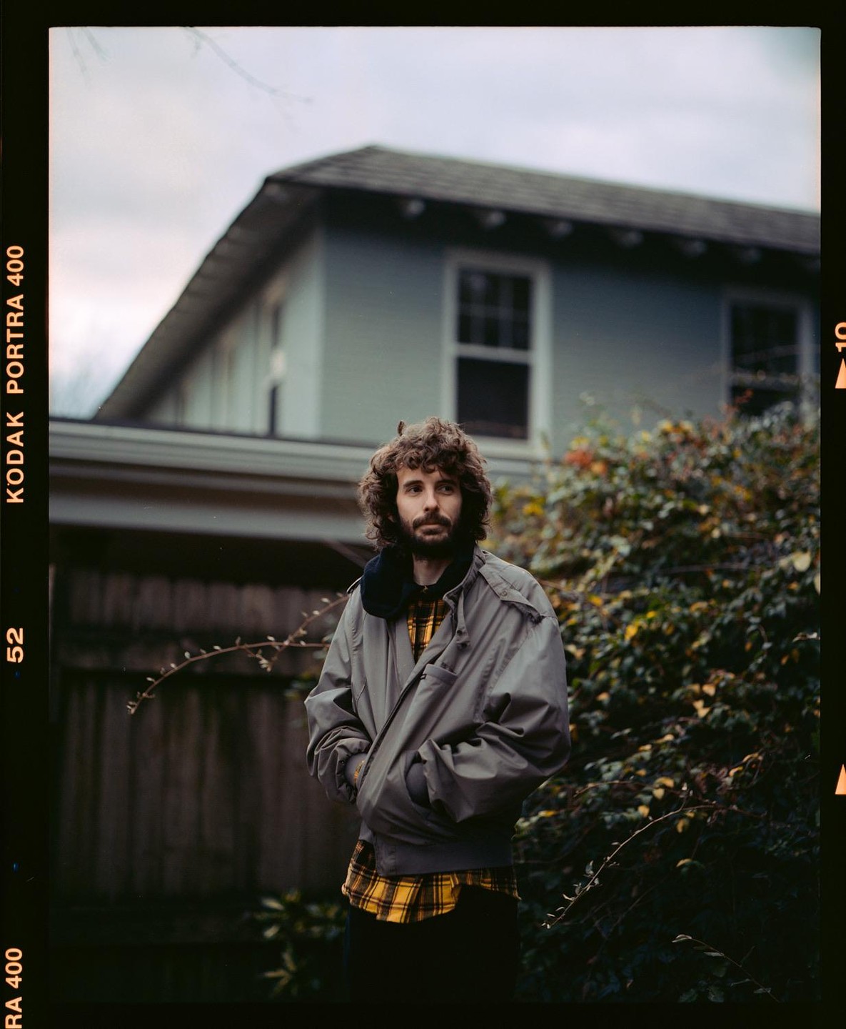 A man with curly hair and a beard stands in front of a house and bushes.