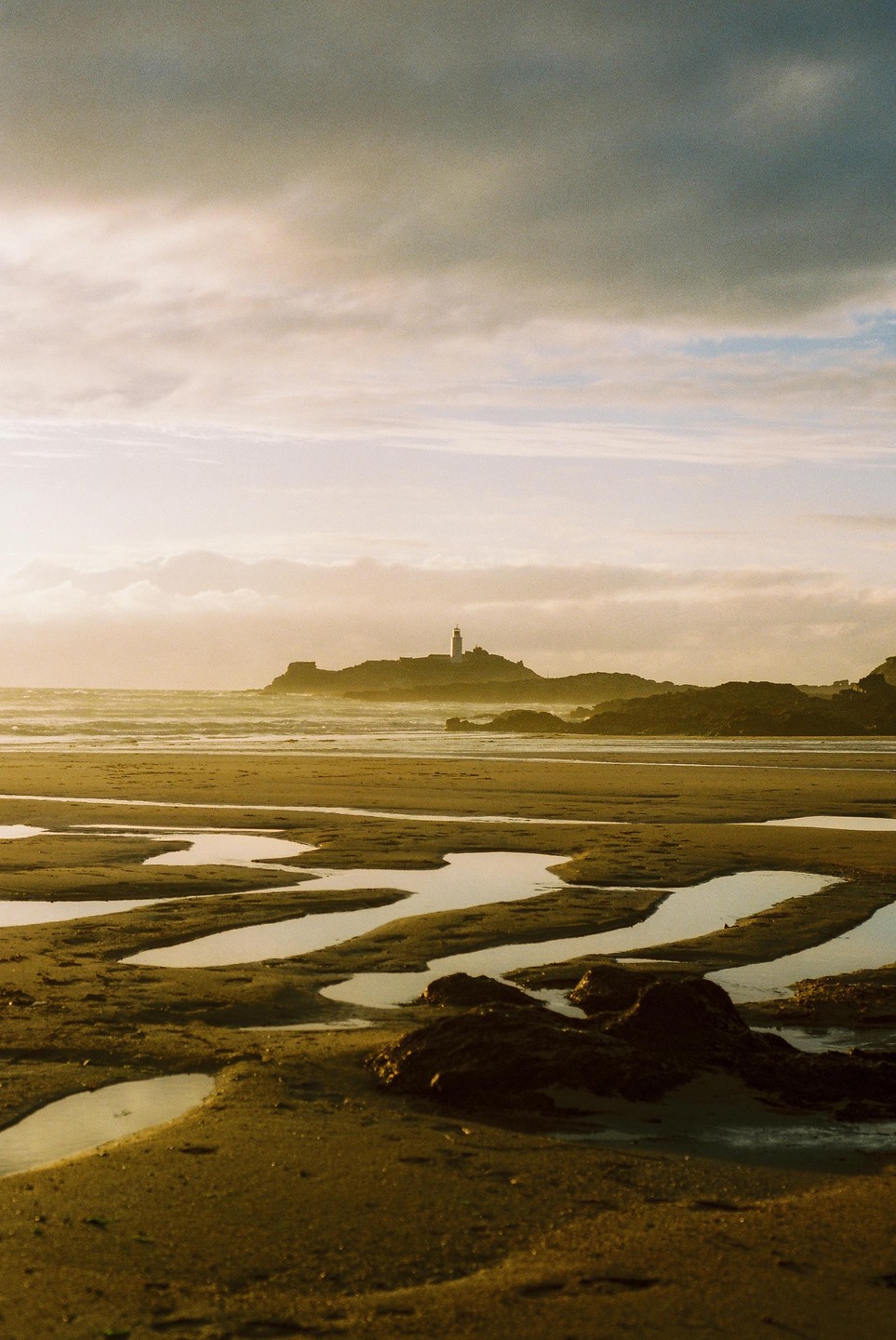 A lighthouse stands on a rocky island in the distance, with a sandy beach and tidal pools in the foreground.