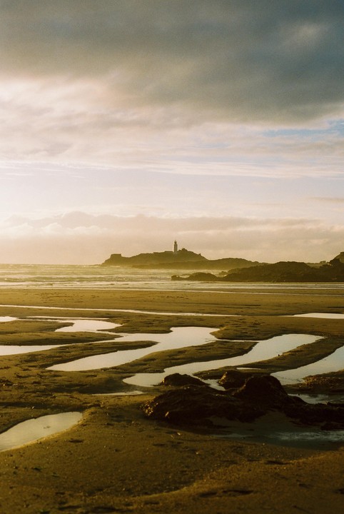 A lighthouse stands on a rocky island in the distance, with a sandy beach and tidal pools in the foreground.