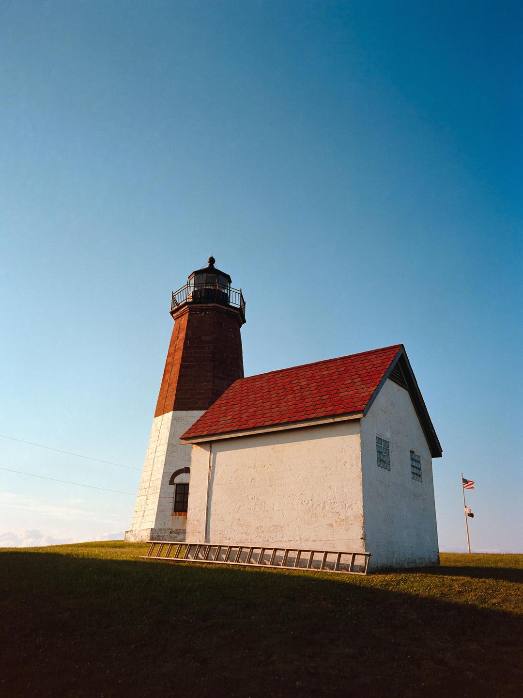 A white and red lighthouse stands on a grassy hill next to a small building with a red roof under a clear blue sky.
